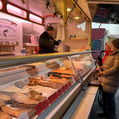 En man klädd i mössa, varm jacka och blått förkläde står i en försäljningsvagn där det säljs fisk. Han betjänar kunder på Ekenäs torg..