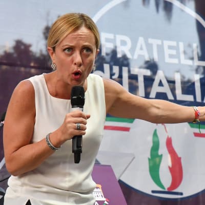 Giorgia Meloni står på en scen med Fratelli d'Italias logga bakom.