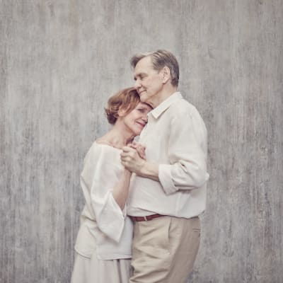 Ett gammalt äkta par står framför en grå vägg och håller om varandra som i en bröllopsvals. 