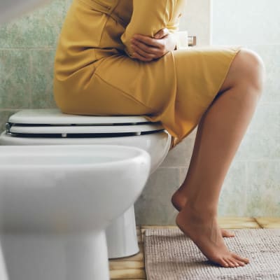 En kvinna i gul klänning som sitter ovanpå en wc-stol och håller sig för magen.