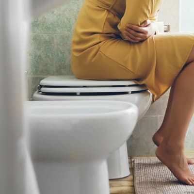 En kvinna i gul klänning som sitter ovanpå en wc-stol och håller sig för magen.