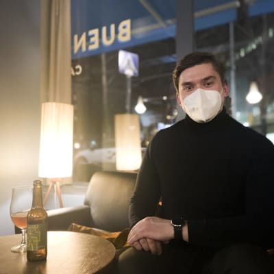 En man ser in i kameran, han har på sig munskydd. Han sitter inne i en bar, på bordet står en ölflaska och ett glas.