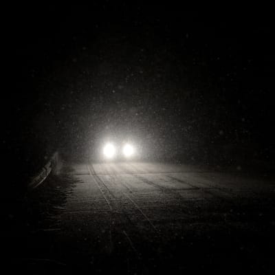 En bil närmar sig på snöig väg i bäcmörker.