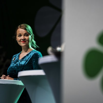 Katri Kulmuni i talarstol med grön fyrklöver på affisch.