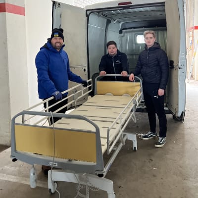 Tre män med en sjukhussäng utanför en skåpbil.