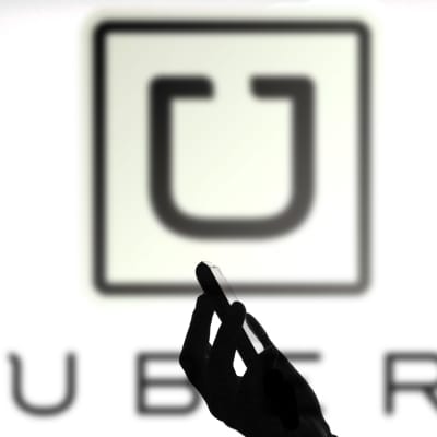I förgrunden en person i profil som tittar på sin smarttelefon, i bakgrunden Ubers logotyp på en vägg. 