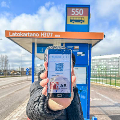 En person visar sin telefon, som visar en nätsida med en bussbiljett. I bakgrunden en busshållplats.