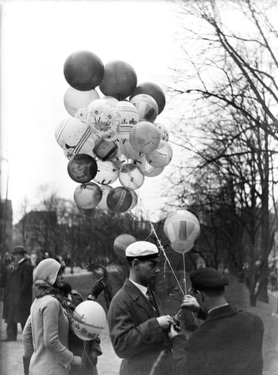 En svartvit gammal bild på en man i studentmössa med ett knippe ballonger i handen. 