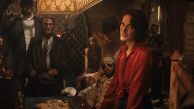 Helena (Phoebe Waller-Bridge) iklädd en röd skjorta och Jürgen Voller (Mads Mikkelsen) i kostym står i ett mörkt rum omgiven av en massa män, de tittar alla åt samma håll och ser allvarliga ut.
