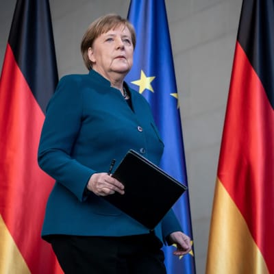 Angela Merkel går på en scen med en mapp i handen. I bakgrunden Tysklands och EU:s flaggor.