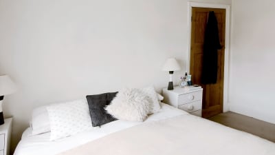 En dubbelsäng som är snyggt bäddad, ljusa färger, vit vägg, nattduksbord med lampa på båda sidorna.