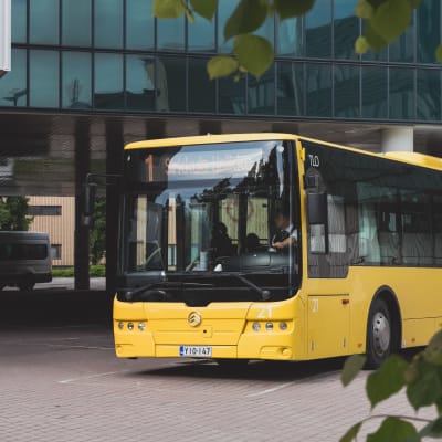 Keltainen bussi kaartaa asemalta kadulle. Kuljettajan vieressä on kaksi matkustajaa. 