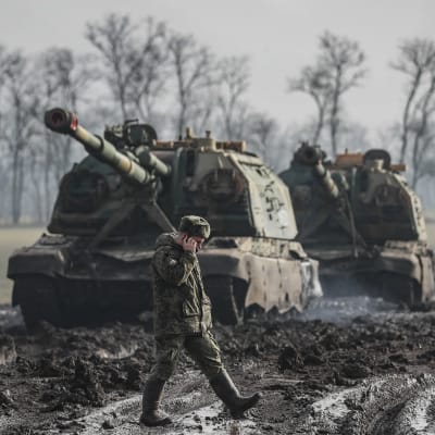 En rysk soldat talar i telefon och vandrar på en lerig väg framför två pansarvagnar.