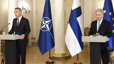 Jens Stoltenberg och Sauli Niinistö står bredvid varandra vid talarpodium och håller tal. I bakgrunden Finlands, Natos och EU:s flagga.