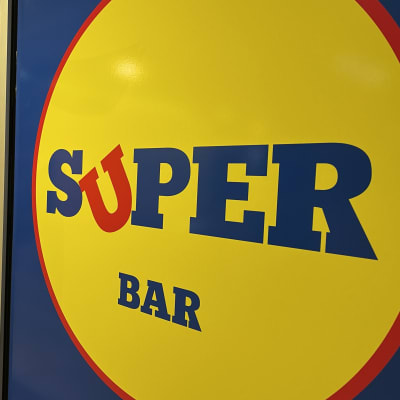 Baarin ulkoseinässä on sinikeltaiset julisteet, joissa on kauppaketju Lidlin logoa muistuttavat Super Barin logot. Alareunassa lukee Drink the pain away.
