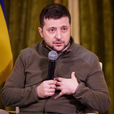 Volodymyr Zelenskyj sitter i en fåtölj. Han tittar åt sidan medan han pratar i en mikrofon som han håller i. I bakgrunden Ukrainas flagga.