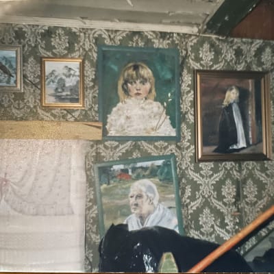 Vanhalla tapetilla päällystetty seinä, johon on ripustettu amatöörin maalaamia muotokuvia.