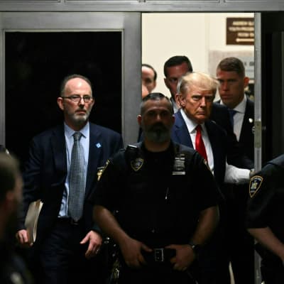 En man i kostym, Donald Trump, stiger in genom en dörr. Framför honom poliser.