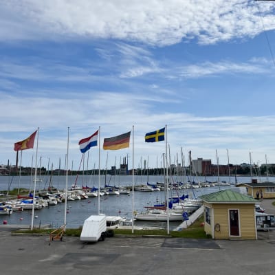 fyra flaggor fladdrar på flaggstänger i vinden. Bakom dem finns bryggor med båtar. I bakgrunden ser man hav och stad. 