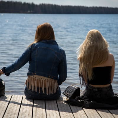 Två tonåriga flickor sitter med ryggen mot kameran på en brygga i sommartid.