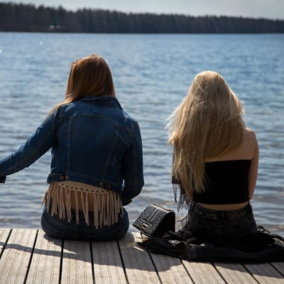 Två tonåriga flickor sitter med ryggen mot kameran på en brygga i sommartid.