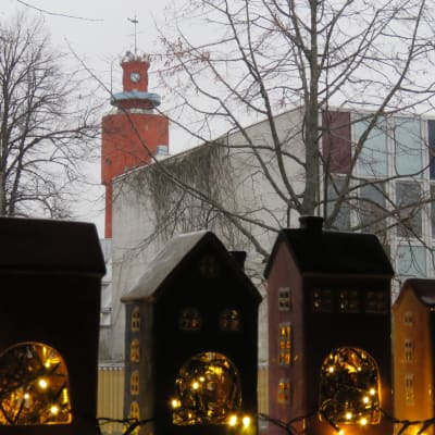 Sex porslinshus uppradadee i ett fönster. Ljusslingor (julljus) inne i husen. Ute kan man se ett gult trähus och Hangös vattentorn.