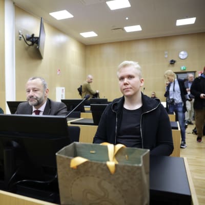 Aleksanteri Kivimäki sitter i en rättssal bredvid sin advokat Peter Jaari. I bakgrunden syns andra människor, bland annat fotografer med kameror.