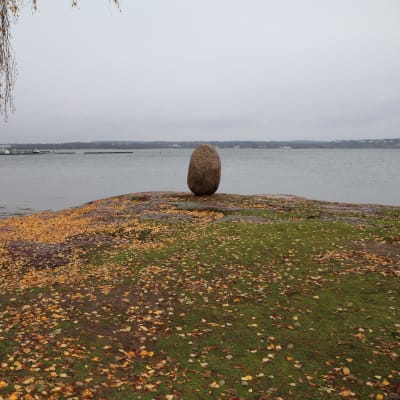 Stenen "Sten Rapakivi" på en klippa med gula höstlöv. I bakgrunden syns en småbåtshamn.