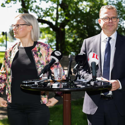 Statsminister Petteri Orpo och finansminister Riikka Purra står framför ett bord med mikrofoner.