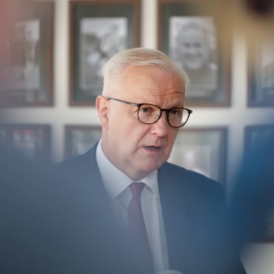 Olli Rehn puhuu. Hänellä on silmälasit, sininen puku, valkoinen kauluspaita ja punapilkullinen kravatti. Taustalla on tauluja.