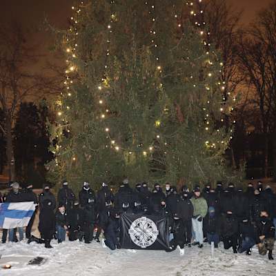 Joukko tummiin pukeutuneita kommandopipoisia henkilöitä poseeraa lippujen kanssa suuren joulukuusen edessä.