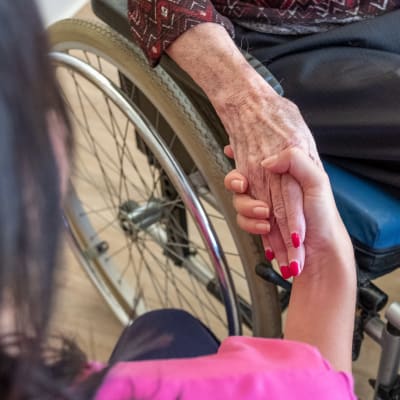 En vårdare håller i en äldre persons hand. Den äldre sitter i rullstol.