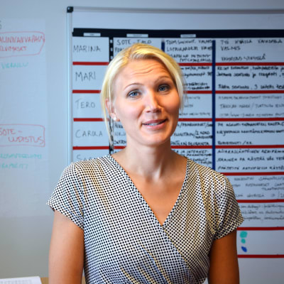 Marina Kinnunen står i sitt kontor. Hon bär en rutig klänning och i bakgrunden syns en whiteboardtavla.