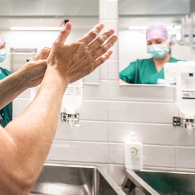 En sjukskötare tvättar händerna i ett badrum. I spegeln syns två sjukskötare som bär ansiktsskydd.