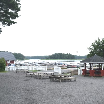 En hamn, i förgrunden syns bord och bänkar och i bakgrunden småbåtar som ligger vid bryggor nära stranden. I bakgrunden öar och vatten.