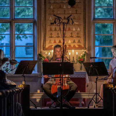 Tre kvinnor sitter och spelar gamla stråkinstrument, barockviolin och gamba, i en kyrka som är upplyst av stearinljus.