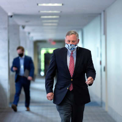 Senaattori Richard Burr kulkee senaatin käytävällä suojainmaski kasvoillaan. Hänellä on tumma puku ja punainen kravatti. Käytävä on vaalea. Kauempana taustalla kulkee toinen mies katsoen puhelintaan.