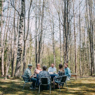 Kuvan etualalla kuusi henkilöä istuu pöydän ääressä ulkosalla kahvilla. Taustalla keväinen koivikko ilman lehtiä.