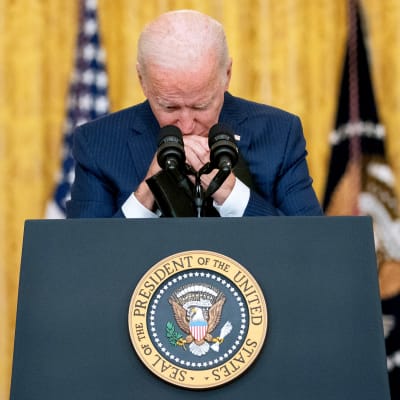 Joe Biden nojaa käsiään puhujanpönttöön ja leukaansa ristissä oleviin käsiinsä. Taustalla näkyy Yhdysvaltain lippu.