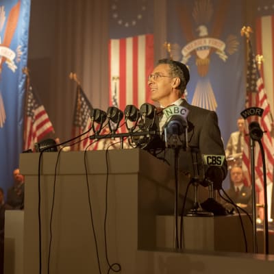 Skådespelaren John Turturro ur en scen från miniserien The Plot Against America. Han står på ett podium inför massa mikrofoner och i bakgrunden syns amerikanska flaggor.