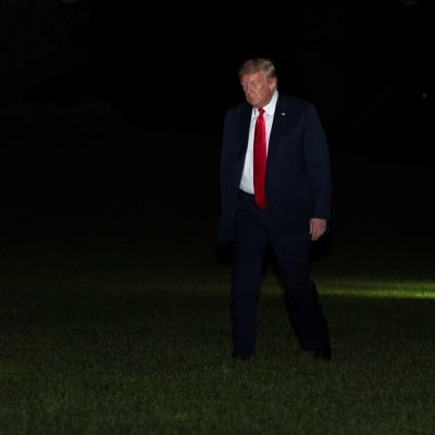 Donald Trump kävelee Valkoisen talon pihanurmella. Tausta on pimeä. Trumpilla on tumma puku ja kirkkaanpunainen kravatti.