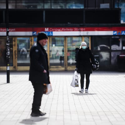 En man i förgrunden och en kvinna med munskydd i bakgrunden som går bort från en metrostation i Helsingfors.