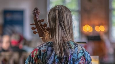 En kvinna i blommig klänning sitter med ryggen åt kameran och spelar på en gamba, ett gammalt stråkinstrument, i en kyrka.