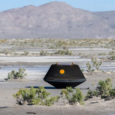 En svart rymdkapsel med en rund orange markering på sidan ligger på marken i ett ökenlandskap med berg i bakgrunden.