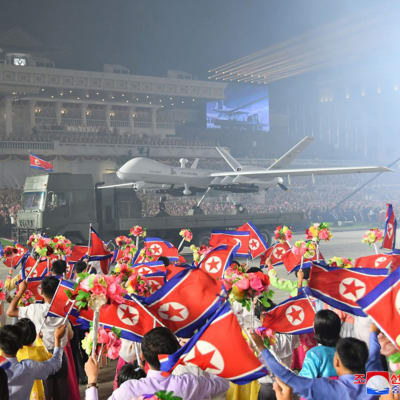 Kuorma-auton lavalla on suurikokoinen drooni, ympärillä suurella aukiolla ihmiset heiluttavat Pohjois-Korean lippuja, ja taustalla näkyy mahtipontinen rakennus.
