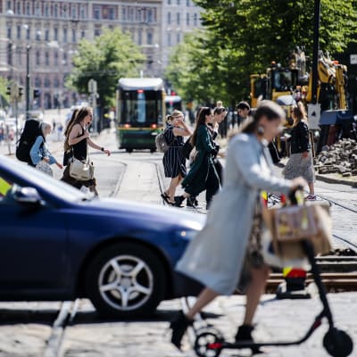 En gatuvy med en blå bil, flera fotgängare en spårvagn och gatuarbeten i Helsingfors centrum i juli 2020.