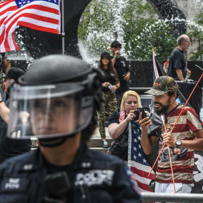 En polis i skyddsutrustning står i förgrunden, i bakgrunden demonstranter som håller i amerikanska flaggan.