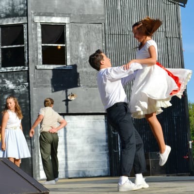 En man lyfter upp en kvinna på en scen i en dans. mannen bär svarta byxor och en skjorta och kvinnan har en vit klänning med rött skärp. De tittar varandra i ögonen. I bakgrunden syns ett högt mörkt hus.