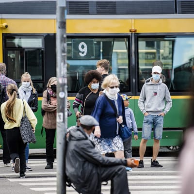Människor vid en spårvagnshålltplats i Helsingfors. En del går över gatan. Många har munskydd på sig.