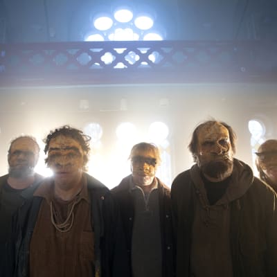 Fem skådespelare med masker i ansiktet står i motljus i en gammal kyrksal.